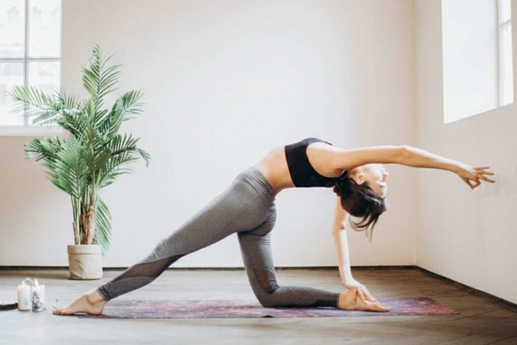 A young brunette woman doing a flexible back stretch yoga pose in a neutral environment with a green leafy plan to the left-hand side background.