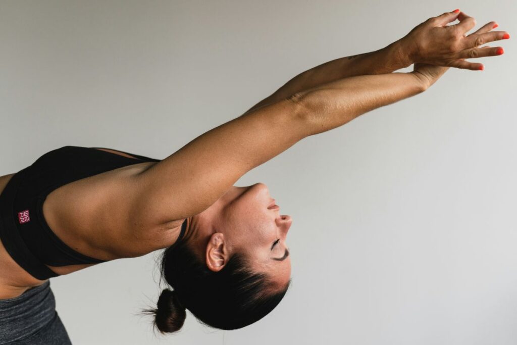 Women with brunette hair in a low bun and black pilates clothes doing a backward stretch with her arms extended.